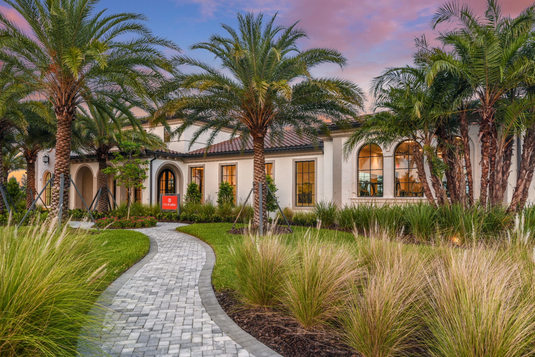 Club Aria resort-style clubhouse with palm-lined paver walk and tropical landscape at dusk