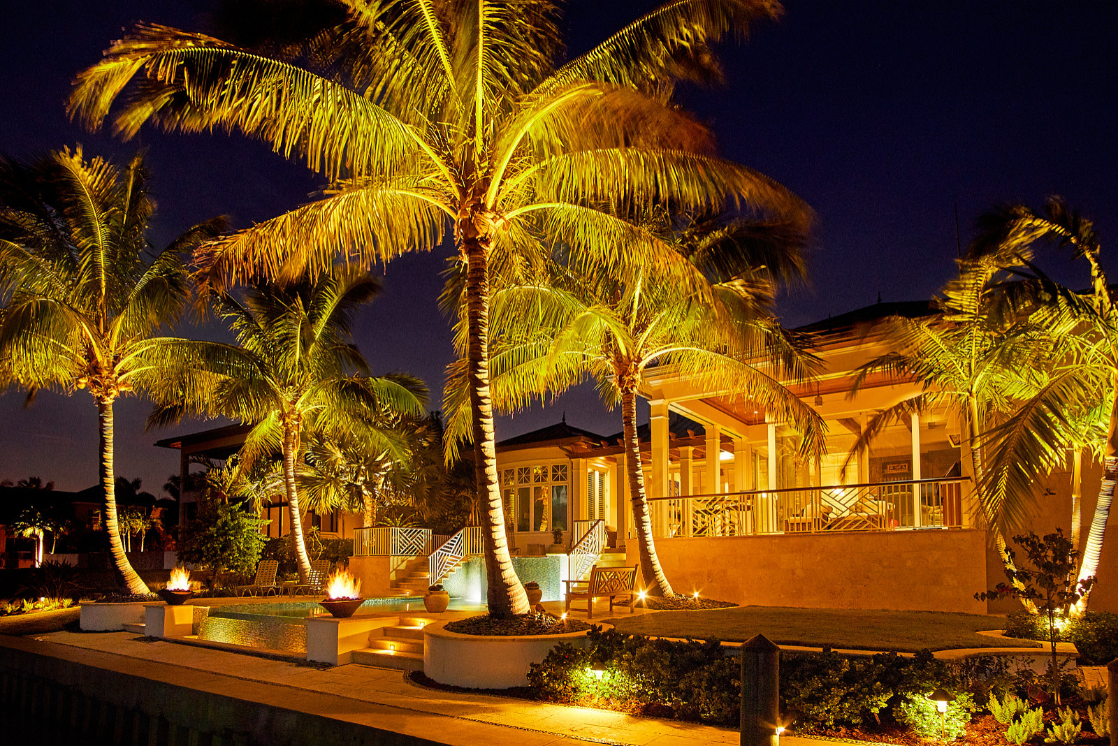 Evening view of a private residence pool with fire bowls, lit palms, and glowing outdoor living space