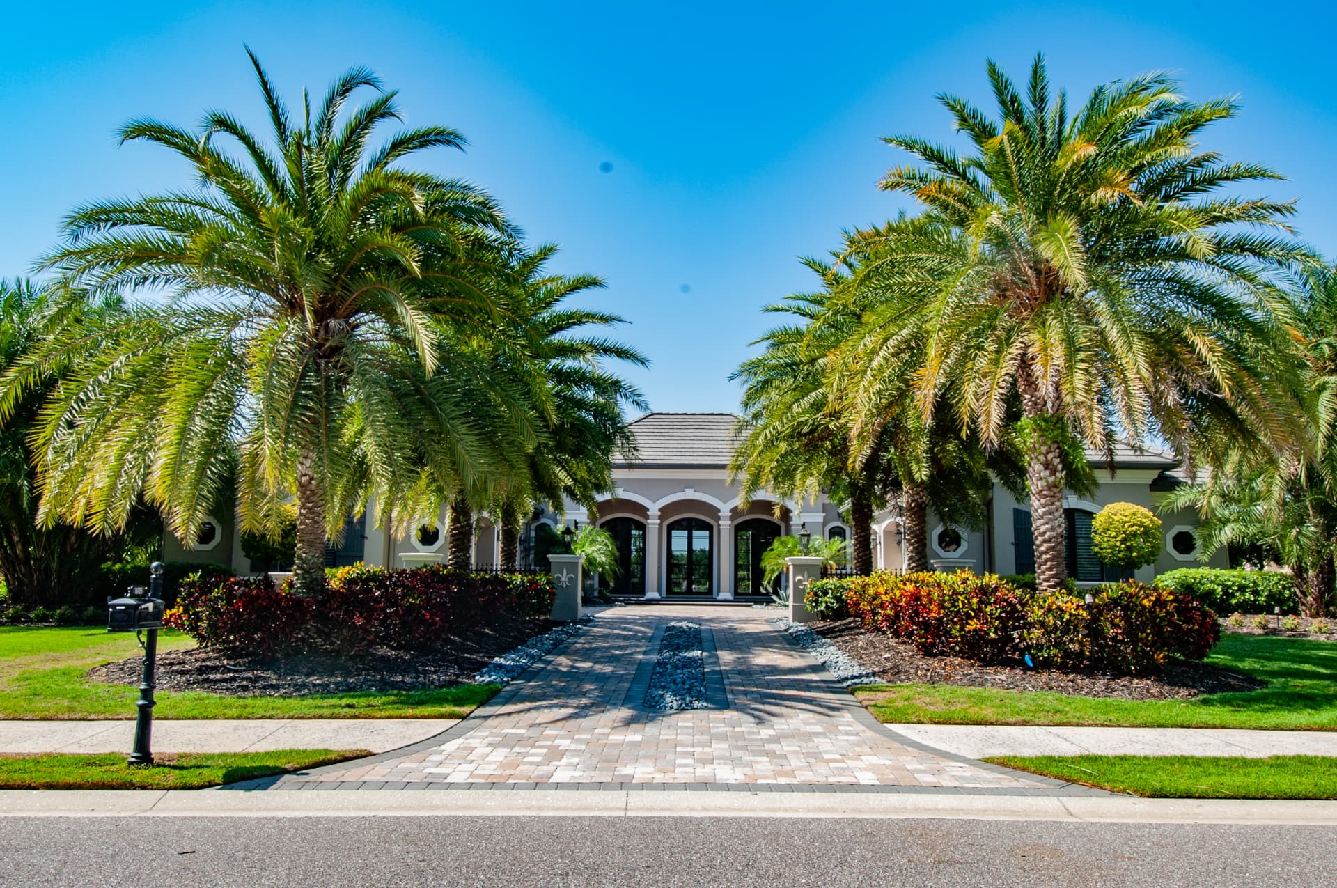 Symmetrical front entrance of a luxury Florida residence designed by a community landscape designer, featuring mature Canary Island palms, colorful garden beds, and a paver driveway under a clear blue sky